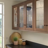 Brown walnut cabinets with glass doors, displaying kitchenware. Below, a tray, plant, and jars on black countertop.