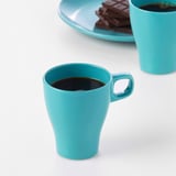 Two turquoise FÄRGRIK mugs, one filled with coffee, sit side by side on a white surface. A plate with chocolate is in the background.