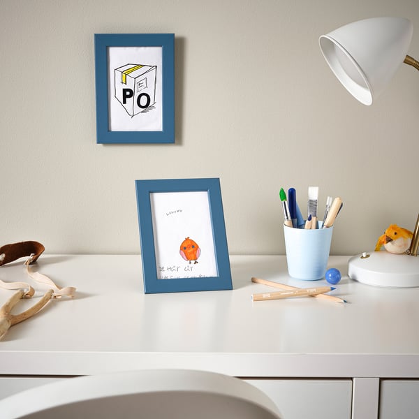 White desk with blue frames, holding drawings, next to a lamp and pen holder.
