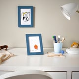 White desk with blue frames, holding drawings, next to a lamp and pen holder.
