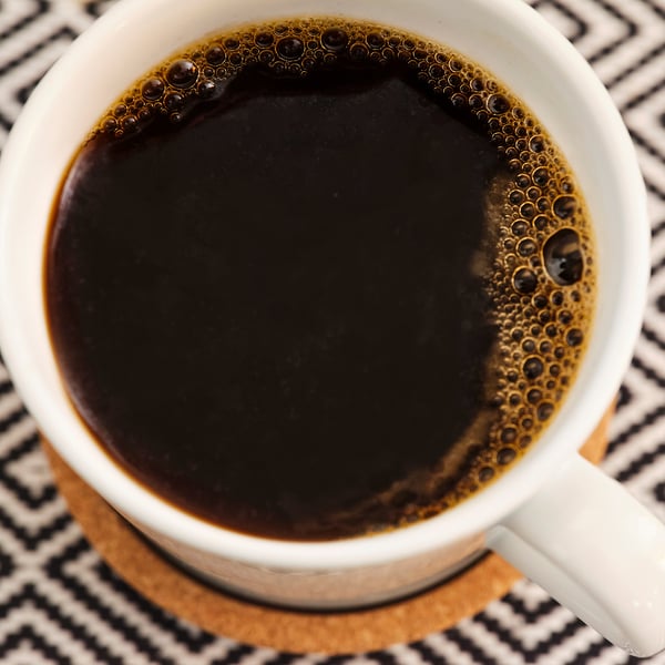 A cup of brewed black coffee, sitting on a patterned coaster. The coffee is dark and bubbly, likely freshly poured.