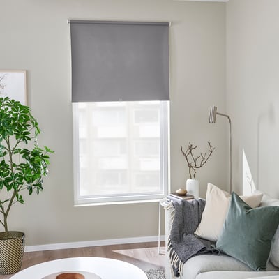 Modern room with grey roller blind, subtle pattern. Cozy chair, pillows, plant, minimalist look.