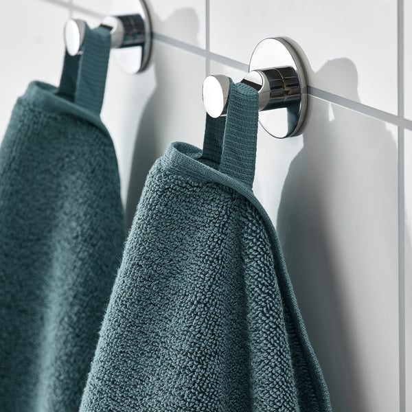 Two green towels hang from a metal rail via hooks. The thick, absorbent towels feature a woven border, showcasing their quality and comfort.