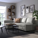 Modern living room with grey convertible sofa, wooden tables, bookshelf, and large window. Sofa is main focus with cushions.