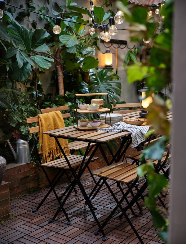 Outdoor dining setup with wooden table, FULLSPÄCKAD bamboo serving tray, string lights, and potted plants.