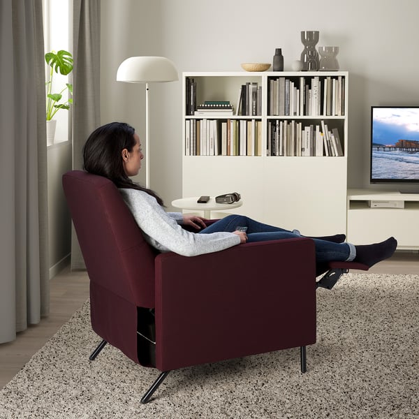 Woman reclines in burgundy chair, remote on side table, cozy living room with tv and bookshelf.