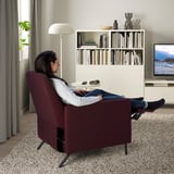 Woman reclines in burgundy chair, remote on side table, cozy living room with tv and bookshelf.
