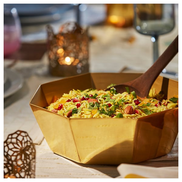 Gold-colored octagonal serving bowl with rice, garnished with red pomegranate seeds and herbs. Wooden spoon in bowl. Placed on beige cloth.