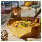 Gold-colored octagonal serving bowl with rice, garnished with red pomegranate seeds and herbs. Wooden spoon in bowl. Placed on beige cloth.