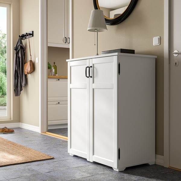 White GREÅKER cabinet stands tall beside door. Holds items behind closed doors, fits well under wall mirror.