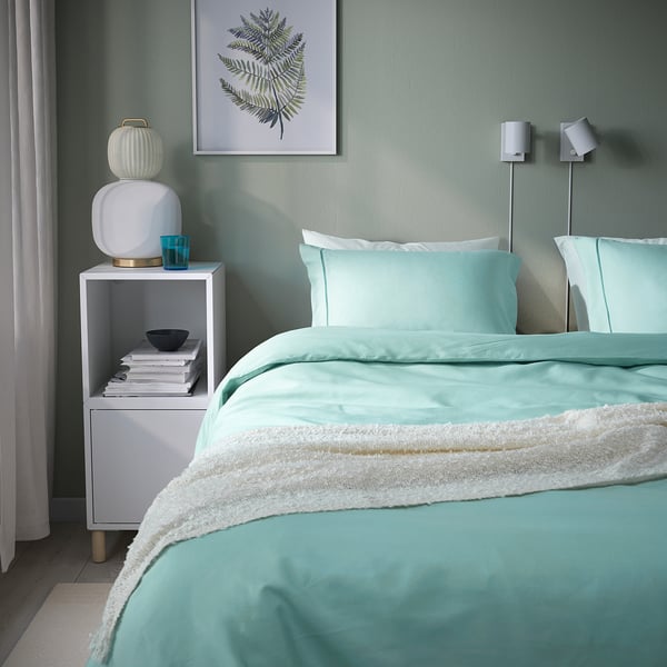 Turquoise duvet set bed with white throw, nightstand with books, fern wall print.