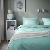 Turquoise duvet set bed with white throw, nightstand with books, fern wall print.