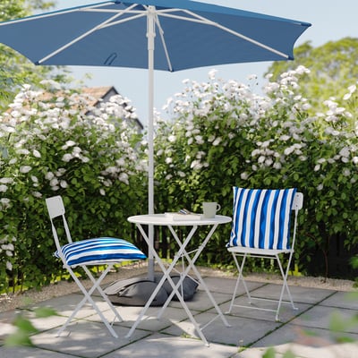 Cozy patio setup with blue-striped cushioned chairs, small table, and large blue umbrella, surrounded by blooming bushes.