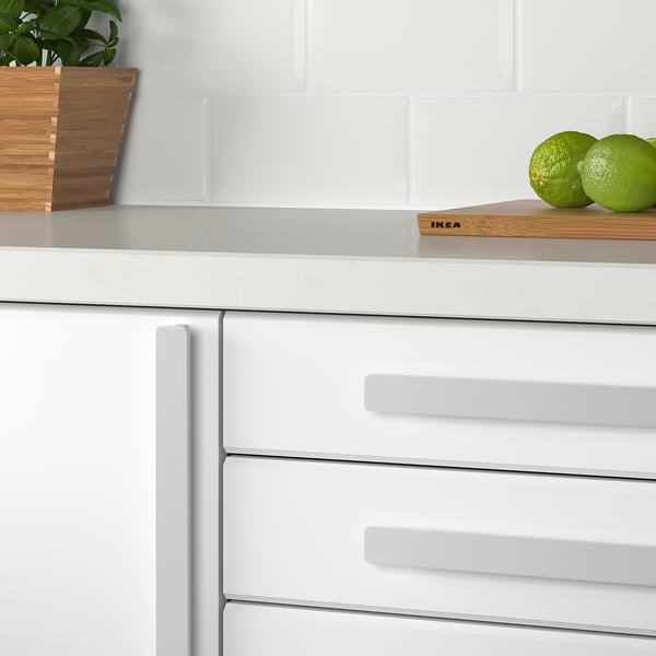 Kitchen with modern white cabinets. Limes on wooden board, plant in basket, gray tile backsplash. Handle labeled ikea.