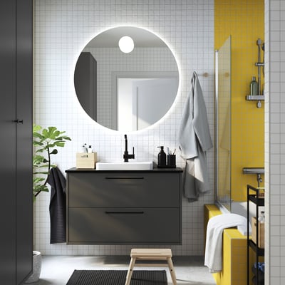 Modern bathroom with round mirror, grey floating vanity, white basin, yellow accent tile, wooden stool, plant, towels, and glass shower.