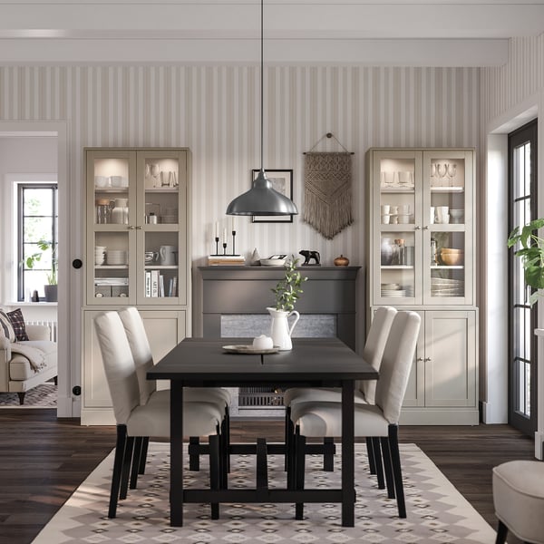 Modern dining room with black HAVSTA table, cream chairs, and striped wallpaper, featuring glass-fronted cabinets displaying whites and greens.