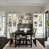 A dining room with a black HAVSTA table, light-colored chairs, and a glass-fronted cabinet.