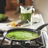 Green pancake batter cooking on a black skillet on stove. Clear mug with green drink and slice of bread nearby.