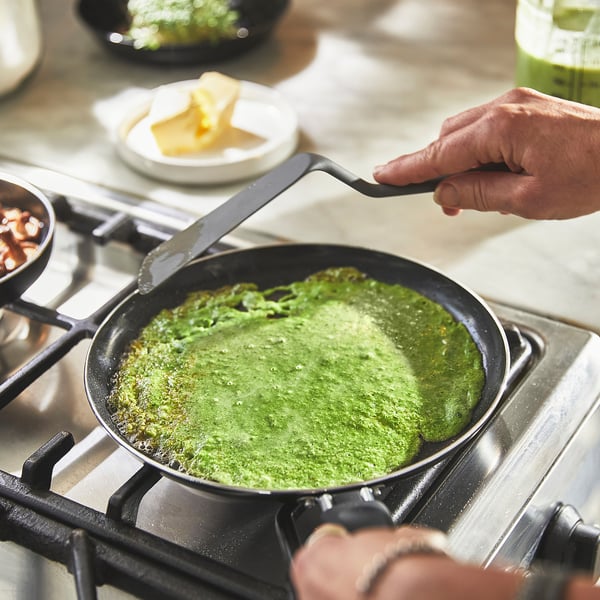 Person is cooking a green pancake in a non-stick frying pan on a stove.
