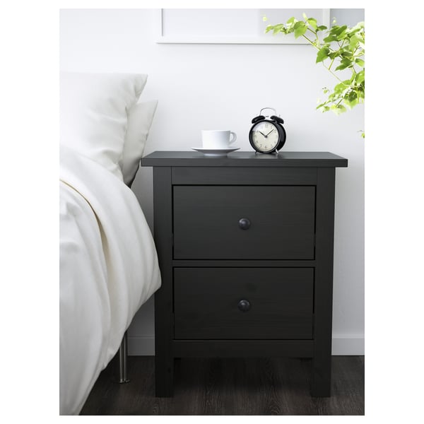 Bedroom setup with HEMNES nightstand. Dark wood, two drawers, smooth handles. On top, a clock and cup. Beside a white bed & green plant.