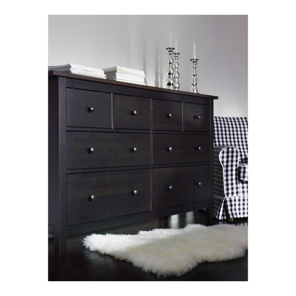 Dark wooden chest of drawers with six drawers. White fur rug is placed on floor. Black and white checkered chair beside. Books and candles on top.