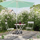 A serene garden scene with a white table, two chairs, a green parasol, and a vase of red flowers.