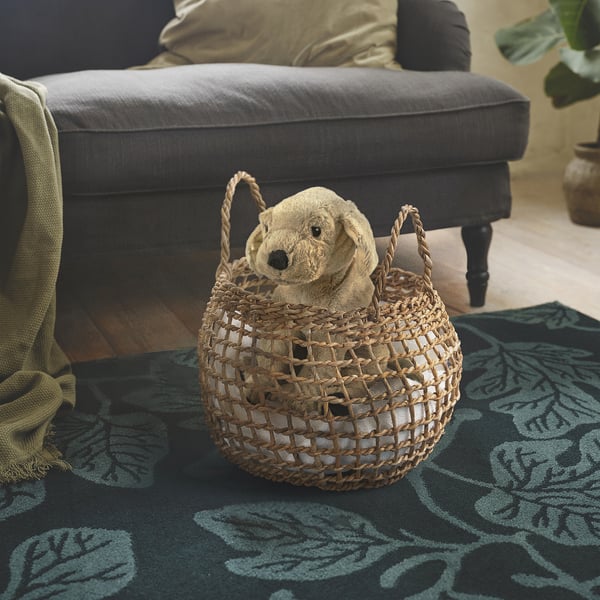 A plush dog toy sits in a woven banana leaf basket with handles, placed on a patterned rug in a cozy, neutral-toned room.