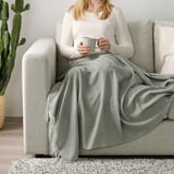 Person sitting on couch, holding mug, wrapped in gray-green throw blanket with fringes.