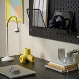 Modern workspace with dark desk, laptop, mouse, stylish lamp, colorful pieces, wall-mounted organizer holding headphones, books, and planter.