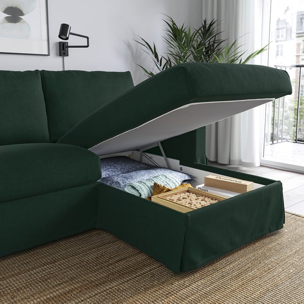 Green sofa with storage ottoman open, revealing blankets and books inside. Modern living room setting with a plant and large window.