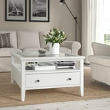 IDANÄS coffee table with glass top & drawer in living room, flanked by sofas, holding vase, books, and bowl.