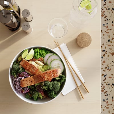 A bowl of salmon, vegetables, and lime slices on a table with chopsticks and a glass of water.