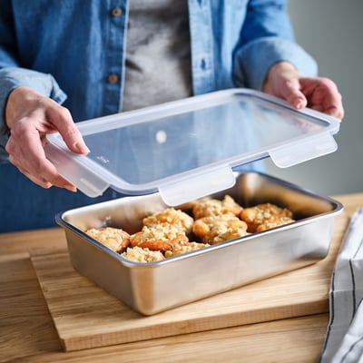 A person placing a transparent lid on a rectangular stainless steel container filled with food, illustrating an easy food storage solution from ikea.