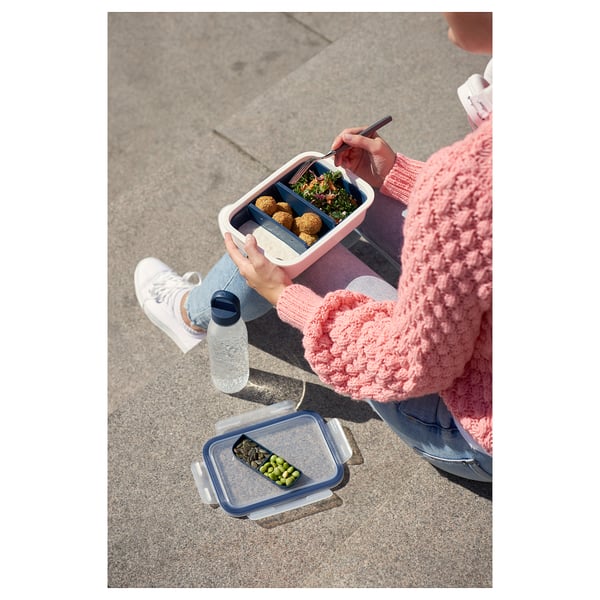 Person in pink sweater holds a divided pink lunchbox with colorful food. Beside them, another empty divided container rests on the ground.