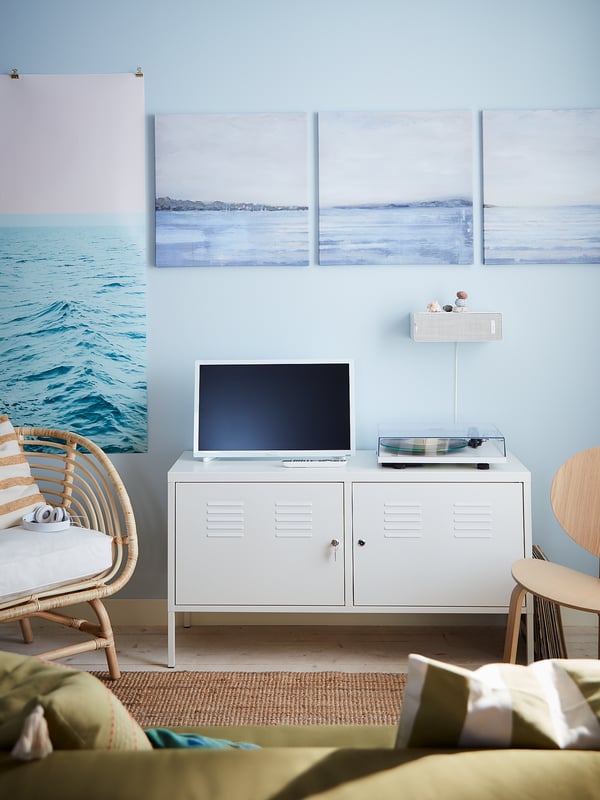 A coastal-themed room with a white IKEA PS metal cabinet featuring a tv, chair, and light blue walls with seascape artwork.
