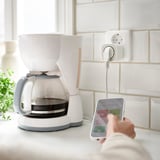A white smart plug connected to a white coffee maker on a kitchen counter.
