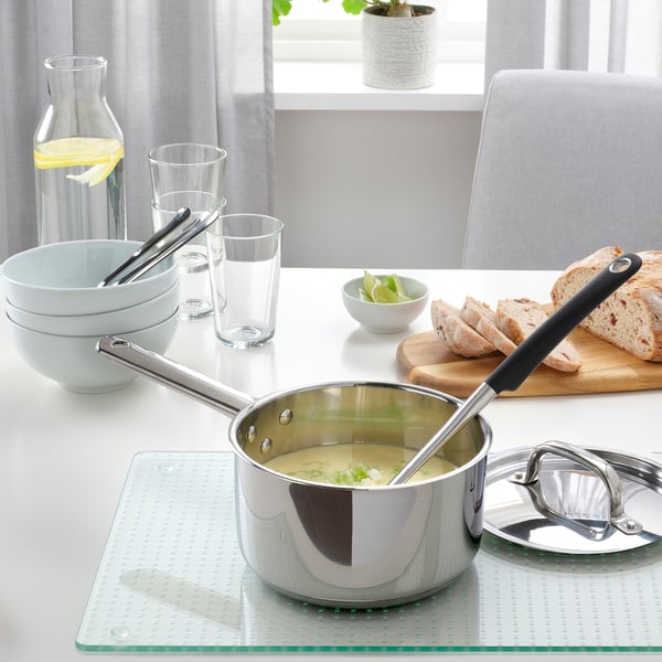 A kitchen table set with a stainless saucepan, bowls, glasses, and bread. The saucepan is on a glass hob protector.