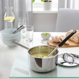 A kitchen table set with a stainless saucepan, bowls, glasses, and bread. The saucepan is on a glass hob protector.
