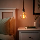 JÄLLBY pendant lamp with warm led over bedside table, multicolored bulb, book, glasses, vase with greenery. Peach wall.