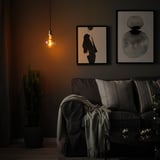 Cozy room with dark grey sofa, grey blanket, hanging warm light bulb, plant, dark artwork & candles.