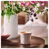 Cozy indoor scene: white ribs vase with pink, purple, white flowers next to a lit gray JÄMNMOD scented candle on wooden table.