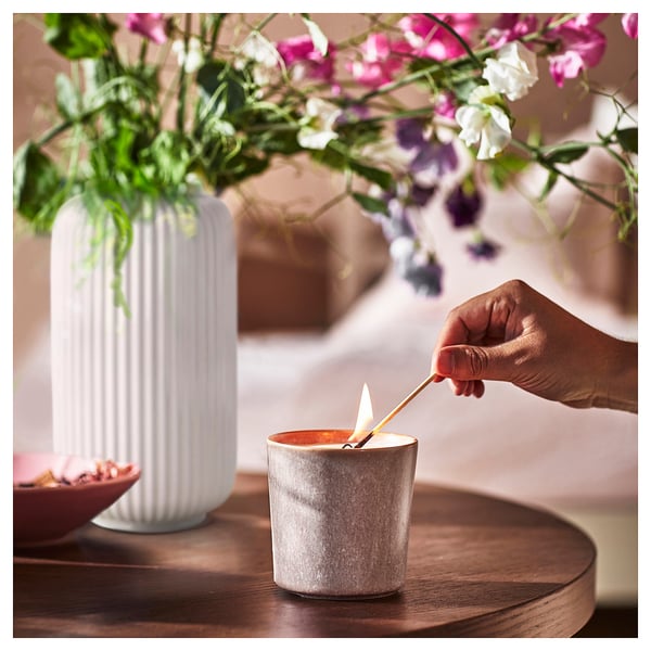 A hand lights a gray JÄMNMOD scented candle on a wooden table, beside a white ribbed vase with purple flowers.