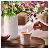 A hand lights a gray JÄMNMOD scented candle on a wooden table, beside a white ribbed vase with purple flowers.