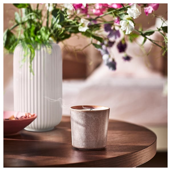 White vase with pink flowers sits next to a gray ceramic candle on a wooden table.