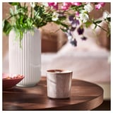 White vase with pink flowers sits next to a gray ceramic candle on a wooden table.
