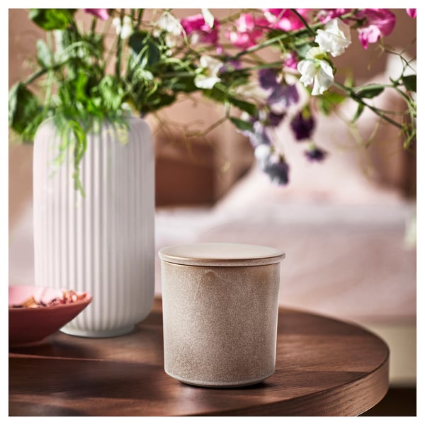 White textured candle in ceramic jar beside ribbed vase with vibrant flowers on wooden table.