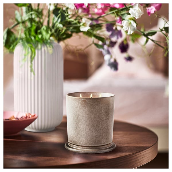 Lit JÄMNMOD candle in beige jar on wooden table with colorful flowers.
