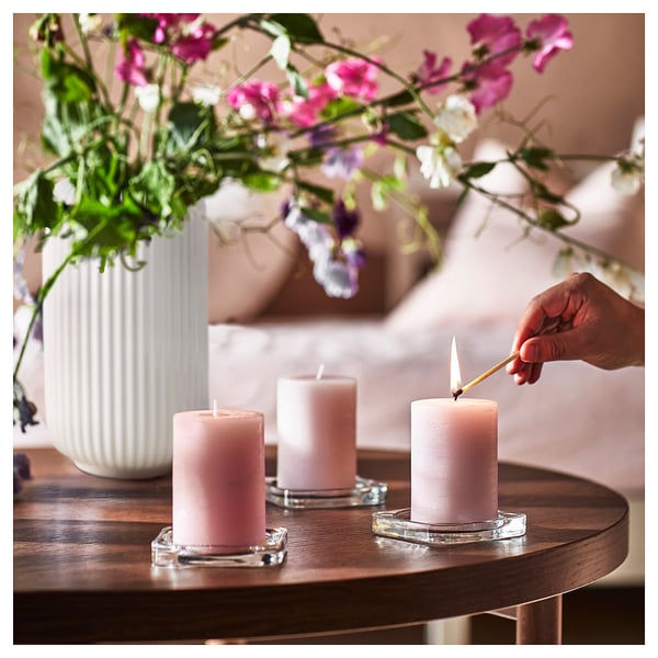 A hand lights three pink JÄMNMOD scented candles on a wooden table. The candles are in glass holders. A white vase with pink flowers is nearby.