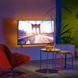 Modern living room with flat screen tv displaying a bridge. Bright red chair, small round tables, and colourful rug with plants.