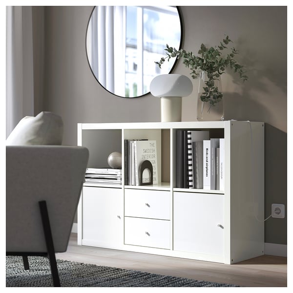 Modern living room with a white ikea KALLAX bookshelf, round mirror, and grey chair. The bookshelf holds books, a lamp, and a vase with greenery.
